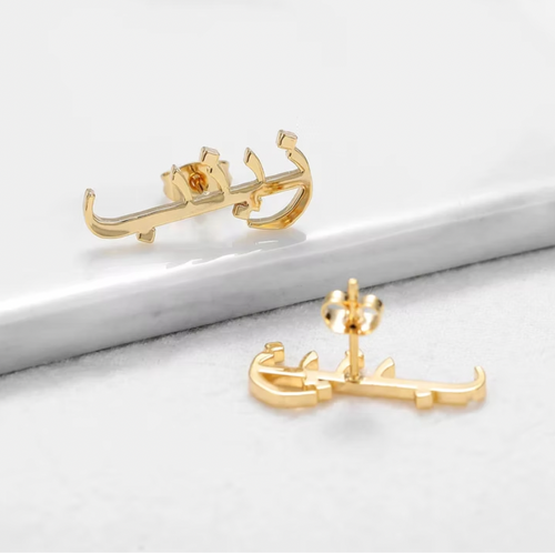 Gold anchor-shaped earrings on a white marble surface. Arabic Name Earr. rose gold. silver plated. silver. gold. silver. rose gold. plated 18k solid sterling silver. makana jewellery jewelry