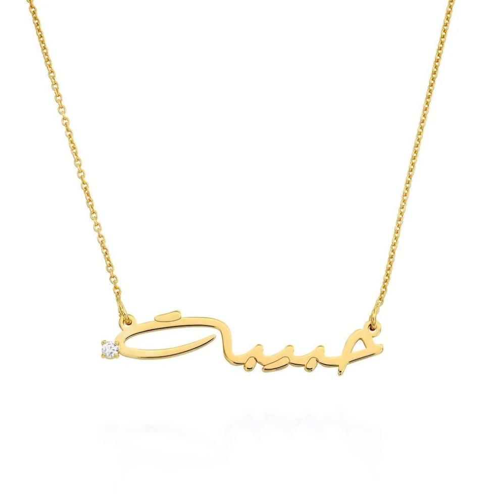 A gold nameplate necklace featuring a cursive script pendant with a small clear gemstone accent on the left side, suspended from a delicate gold chain. Diamond, sterling silver 925, gold plated, rose gold plated. makana jewellery, jewelry.